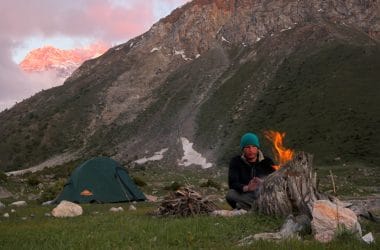 Man sitting beside a campfire with tent and mountains in background