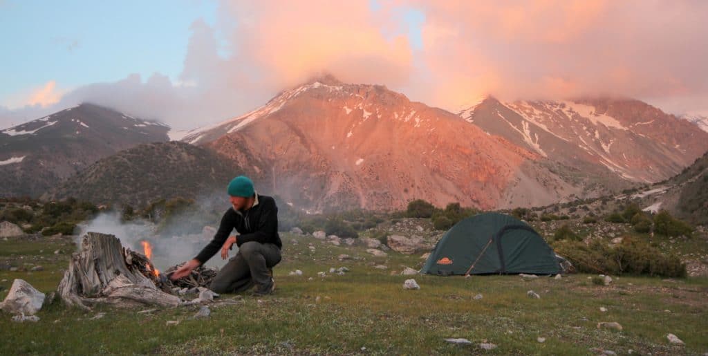 Male hiker tending a fire with tent and mountains in background
